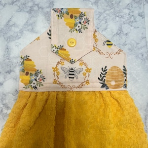May include: A yellow kitchen towel with a decorative top featuring a bee and honeycomb design. The top has a beige background with floral and bee illustrations, and a yellow button. The towel is made of a textured, absorbent material.