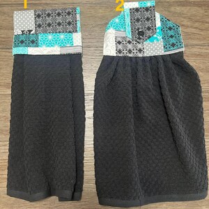 May include: Two hanging kitchen towels. The towels are black with a textured pattern. The top portion of each towel features a patchwork design with teal, gray, white, and black patterned fabric. One towel has a straight top, and the other has a button closure.