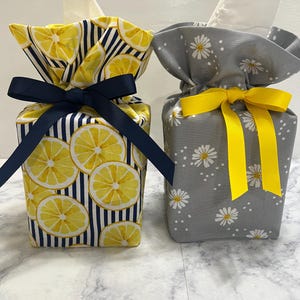 May include: Two fabric tissue box covers. The left cover features a yellow lemon and navy blue stripe pattern with a navy blue ribbon bow. The right cover features a grey background with white daisy flowers and a bright yellow ribbon bow.