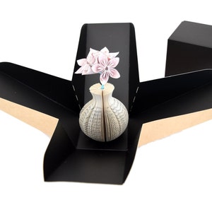 K&ouml;nnte beinhalten: Eine schwarze Origami-Box mit braunem Innenraum, ge&ouml;ffnet, um eine wei&szlig;e Buchvase mit einem rosafarbenen Origami-Blumenarrangement zu enth&uuml;llen.