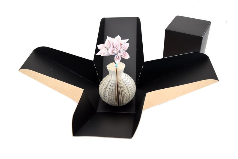 K&ouml;nnte beinhalten: Eine schwarze Origami-Box mit braunem Innenraum, ge&ouml;ffnet, um eine wei&szlig;e Buchvase mit einem rosafarbenen Origami-Blumenarrangement zu enth&uuml;llen.