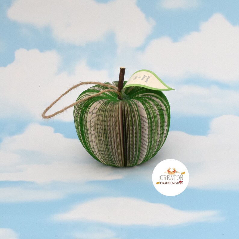 May include: A green book-shaped apple ornament with a brown twig stem and a small tag that reads "The Journey of a Thousand Miles Begins with a Single Step".