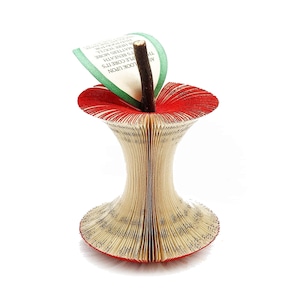 May include: A red and green paper apple sculpture made from the pages of a book. The pages are folded to create the shape of an apple, and the stem is made from a twig. The text on the pages is visible, and the words "LOOK UPON THE CORE IT'S AS T'S BENEATH MATTERS MORE OR HERE YOU'L FIND FOOD FOR" are visible.