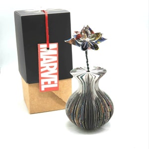 以下が含まれることがあります： 「MARVEL」と赤いタグの付いた黒い箱が、折り畳まれたコミックでできた花瓶の隣にあります。白い真珠の中心を持つ紙の花が花瓶に入っています。