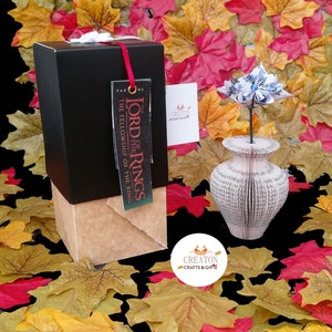 May include: A black gift box with a brown paper wrapping and a tag that reads "The Lord of the Rings The Fellowship of the Ring". A blue paper flower is in a white book vase. The background is a pile of fall leaves.