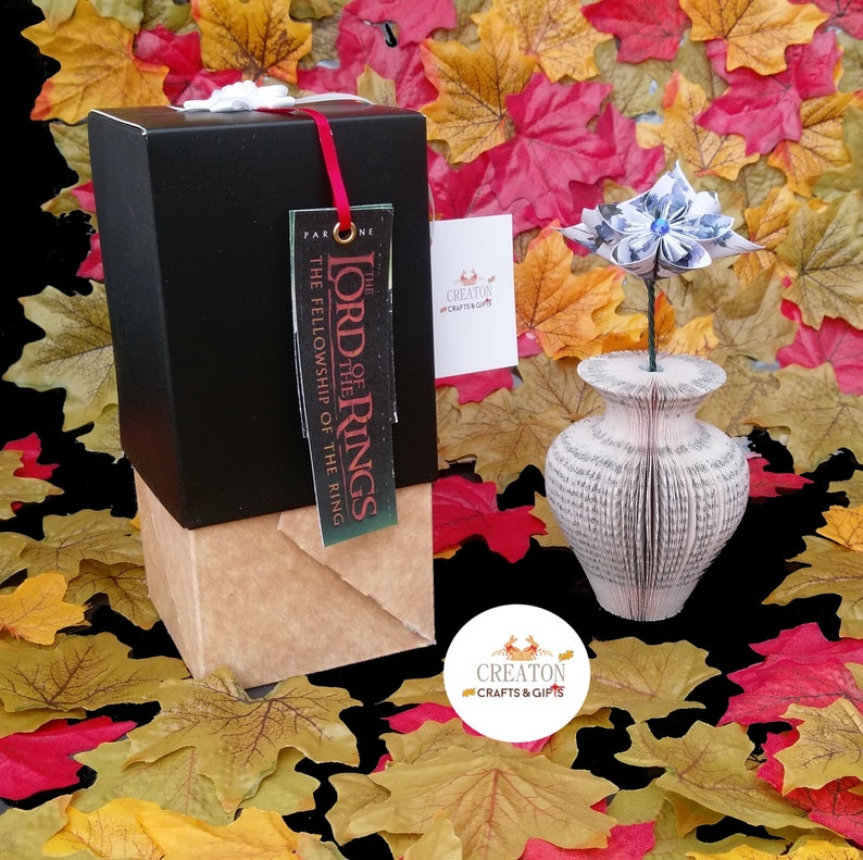 May include: A black gift box with a brown paper wrapping and a tag that reads "The Lord of the Rings The Fellowship of the Ring". A blue paper flower is in a white book vase. The background is a pile of fall leaves.