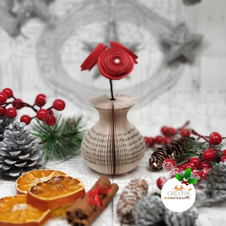 K&ouml;nnte beinhalten: Eine rote Papierblumen-Arrangement in einer Vase aus einem gefalteten Buch. Die Vase ist umgeben von Dekoartikeln, darunter Zimtstangen, getrocknete Orangenscheiben und Tannenzapfen.