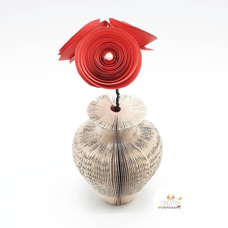K&ouml;nnte beinhalten: Eine rote Papierblumen-Arrangement in einer Vase aus den Seiten eines Buches. Die Vase ist wei&szlig; und hat eine strukturierte Oberfl&auml;che. Die Blume besteht aus mehreren Papierschichten und hat eine Perle in der Mitte.