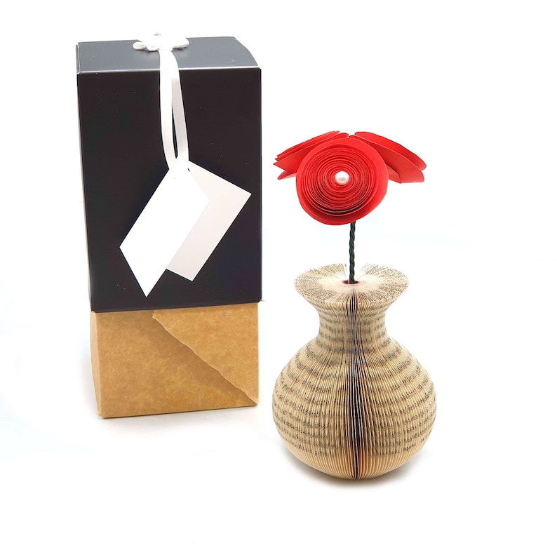 K&ouml;nnte beinhalten: Eine rote Papierblume in einer Vase aus den Seiten eines Buches. Die Vase steht auf einem Tisch neben einer braunen Geschenkbox mit einem wei&szlig;en Etikett.