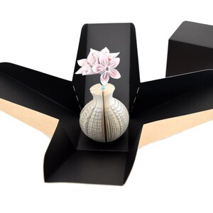 May include: A black origami box with a brown interior, open to reveal a white book vase with pink origami flowers.