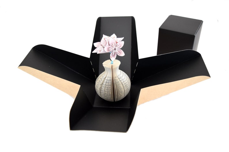 May include: A black origami box with a brown interior, open to reveal a white book vase with pink origami flowers.