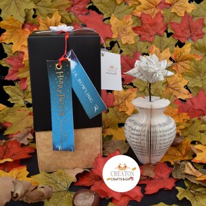 May include: A black gift box with a blue and white Harry Potter book tag and a white flower made from the pages of a book in a white vase. The vase is also made from the pages of a book. The background is a pile of fall leaves.