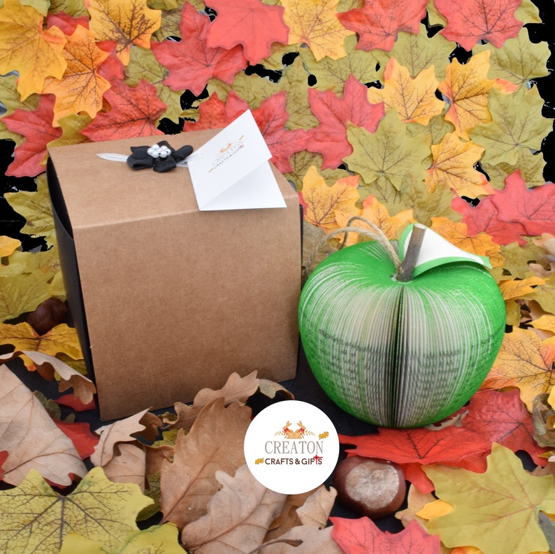 May include: A green book shaped like an apple, with a brown box and a black bow on top. The box has a white tag with the text "Creaton Crafts & Gifts". The apple and box are surrounded by fall leaves.