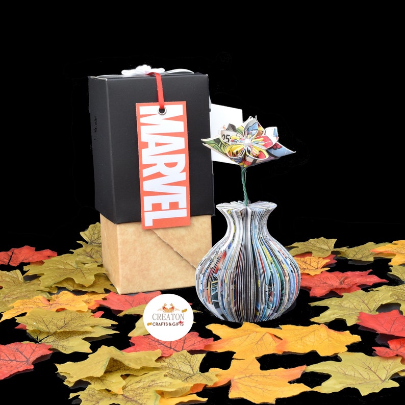 May include: A black box with a red "MARVEL" tag and a brown paper wrapping. A vase made from comic books with a flower made from comic book pages is in front of the box. The vase and flower are on a bed of fall leaves.