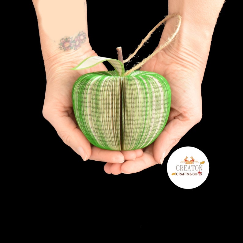 May include: A green and white book-shaped apple ornament, folded from the pages of a book, held in two hands. The ornament has a green stem and leaf. The ornament is labeled "Creaton Crafts & Gifts".