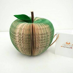 May include: A green book-shaped apple sculpture made from the pages of a book. The apple has a green leaf on top and is sitting on a white surface. The text "Creaton Crafts & Gifts" is visible on a white tag.