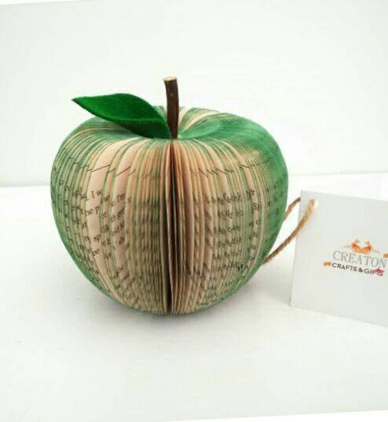 May include: A green book-shaped apple sculpture made from the pages of a book. The apple has a green leaf on top and is sitting on a white surface. The text "Creaton Crafts & Gifts" is visible on a white tag.