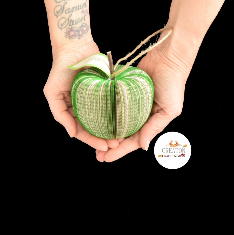 May include: A green book-shaped apple ornament made from pages of a book. The apple is being held in the hands of a person. The ornament has a string attached to the top for hanging.