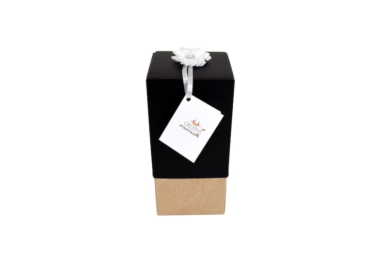May include: A black gift box with a brown base, tied with a white ribbon and a white flower. The box has a white tag with the text "CREATION CRAFTS & GIFTS".