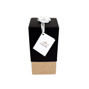 K&ouml;nnte beinhalten: Eine schwarze Geschenkbox mit braunem Boden, gebunden mit einem wei&szlig;en Band und einer wei&szlig;en Blume. Die Box hat ein Etikett mit dem Text "CREATION CRAFTS & GIFTS".