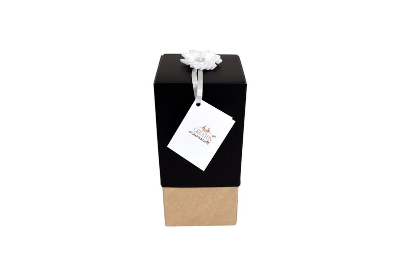 K&ouml;nnte beinhalten: Eine schwarze Geschenkbox mit braunem Boden, gebunden mit einem wei&szlig;en Band und einer wei&szlig;en Blume. Die Box hat ein Etikett mit dem Text "CREATION CRAFTS & GIFTS".