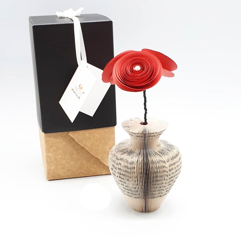 K&ouml;nnte beinhalten: Eine rote Papierblume in einer Vase aus den Seiten eines Buches. Die Vase ist hellbraun und die Blume besteht aus mehreren Schichten roten Papiers.
