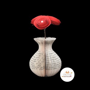 K&ouml;nnte beinhalten: Eine rote Papierblumen-Arrangement in einer Vase aus den Seiten eines Buches. Die Vase ist wei&szlig; und hat eine strukturierte Oberfl&auml;che. Das Blumenarrangement besteht aus drei roten Papierblumen.