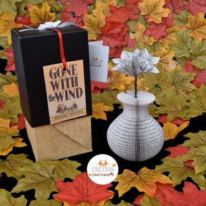 May include: A book vase with a white paper flower arrangement. The vase is made from the pages of the book "Gone With The Wind" by Margaret Mitchell. The vase is sitting on a bed of fall colored leaves.