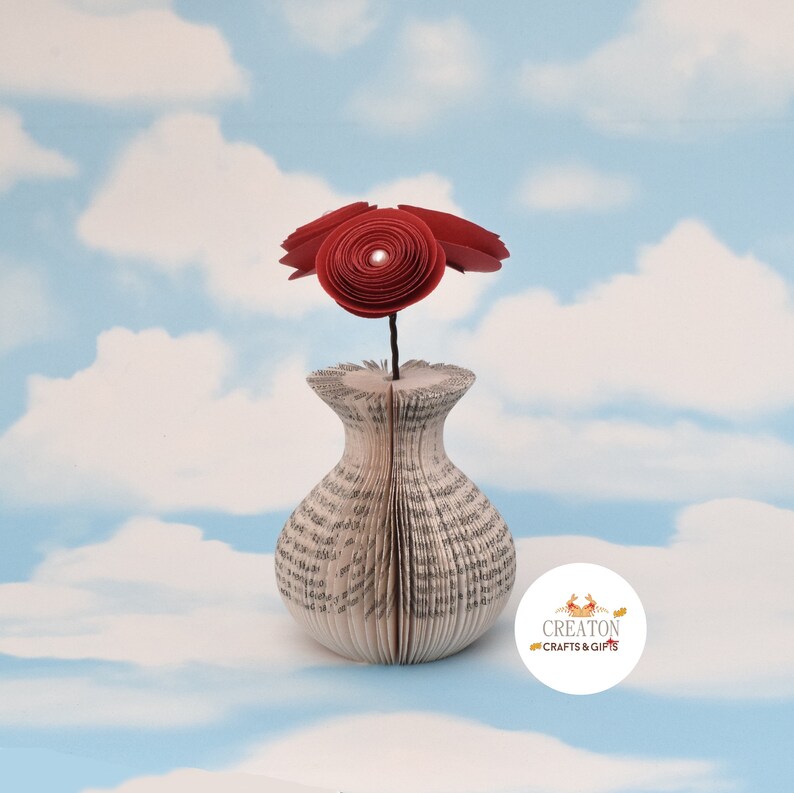 K&ouml;nnte beinhalten: Eine rote Papierblumen-Arrangement in einer Vase aus den Seiten eines Buches. Die Vase ist eine einzigartige und kreative M&ouml;glichkeit, Blumen zu pr&auml;sentieren.