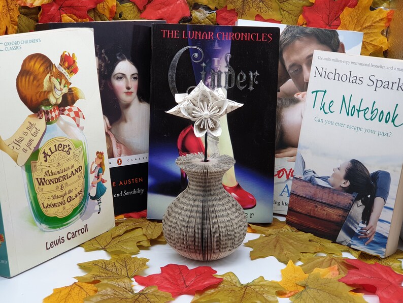 May include: A white paper flower in a vase made from the pages of a book. The vase is sitting on a table with several fall leaves around it. There are four books in the background, including "Alice's Adventures in Wonderland" by Lewis Carroll, "Pride and Prejudice" by Jane Austen, "The Lunar Chronicles" by Marissa Meyer, and "The Notebook" by Nicholas Sparks.