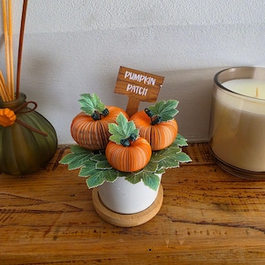May include: A white ceramic pot with a wooden base containing three orange paper pumpkins and green paper leaves. A brown wooden sign with the white text PUMPKIN PATCH is inserted into the centre. The pumpkins feature thin vertical paper folds.