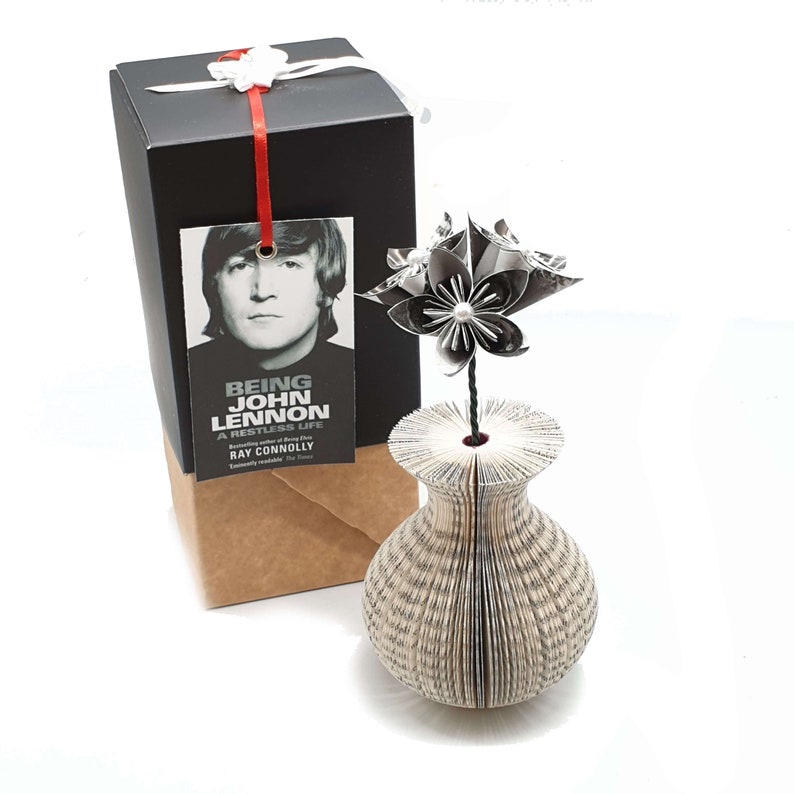 May include: A black and white origami flower in a book vase. The vase is made from the pages of a book titled "Being John Lennon: A Restless Life" by Ray Connolly.