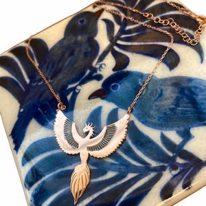May include: A rose gold phoenix pendant necklace. The pendant features a stylized phoenix with outstretched wings and a flowing tail. The necklace is displayed on a blue and white patterned surface.