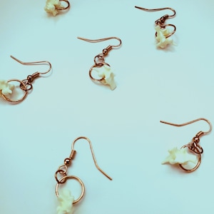 May include: Copper-colored wire earrings with a small, off-white bone-shaped charm. The earrings have a simple hook design and a small circular accent. The bone charms are clustered around the circular accent.