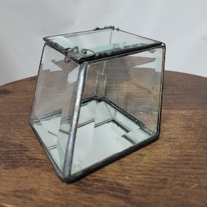 May include: A small, clear glass box with a hinged lid, constructed with dark, soldered metal edges. The box features a trapezoidal side profile and a reflective mirrored base, showcasing a clean, geometric design with visible metal seams.