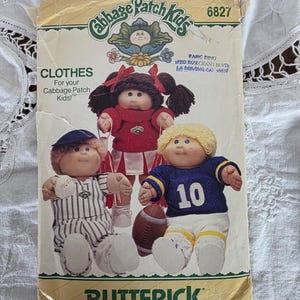 May include: Vintage Butterick pattern for Cabbage Patch Kids clothes. The pattern features illustrations of three Cabbage Patch Kids dolls in various outfits, including a cheerleader, baseball player, and football player. The pattern number is 6827.
