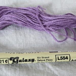 May include: A skein of lavender-colored Galaxy Deluxe Floss, six-strand embroidery thread. The label reads "L554 Art. 711" and includes the price "21¢". The thread is loosely arranged, showing its texture and color. Ideal for embroidery and cross-stitch projects.
