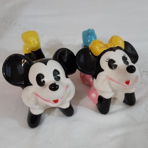 Może przedstawiać: Para ceramicznych figurek Myszki Miki i Myszki Minnie. Miki ma czarne uszy, ż&oacute;łte rękawice i czarne buty. Myszka Minnie ma ż&oacute;łtą kokardę, r&oacute;żową sp&oacute;dnicę w kropki i czarne buty. Oboje mają białe twarze i czerwone uśmiechy.