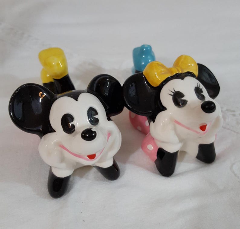 Może przedstawiać: Para ceramicznych figurek Myszki Miki i Myszki Minnie. Miki ma czarne uszy, ż&oacute;łte rękawice i czarne buty. Myszka Minnie ma ż&oacute;łtą kokardę, r&oacute;żową sp&oacute;dnicę w kropki i czarne buty. Oboje mają białe twarze i czerwone uśmiechy.