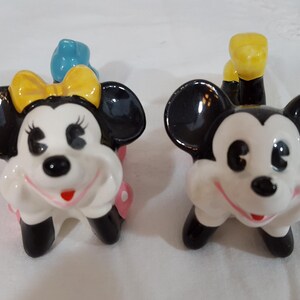 Może przedstawiać: Para ceramicznych figurek przedstawiających Myszki Miki i Minnie. Miki ma czarne uszy i ż&oacute;łty ogon. Myszka Minnie ma ż&oacute;łtą kokardę i niebieską kokardę na głowie. Oboje mają białe twarze i czerwone uśmiechy.