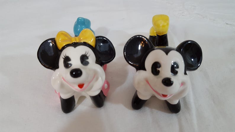 Może przedstawiać: Para ceramicznych figurek przedstawiających Myszki Miki i Minnie. Miki ma czarne uszy i ż&oacute;łty ogon. Myszka Minnie ma ż&oacute;łtą kokardę i niebieską kokardę na głowie. Oboje mają białe twarze i czerwone uśmiechy.