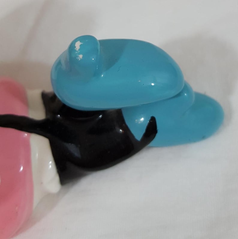 Może przedstawiać: Ceramiczna figurka z jasnoniebieskim kapeluszem, czarną kurtką i r&oacute;żowym tułowiem. Figurka ma błyszczące wykończenie i jest umieszczona na białym tle. Kapelusz ma mały odprysk.