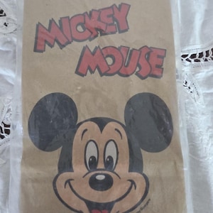 May include: A brown paper bag featuring a smiling Mickey Mouse face and the words "Mickey Mouse" in red letters. The bag has black mouse ears and a red mouth. The text "The Walt Disney Company" is printed at the bottom.