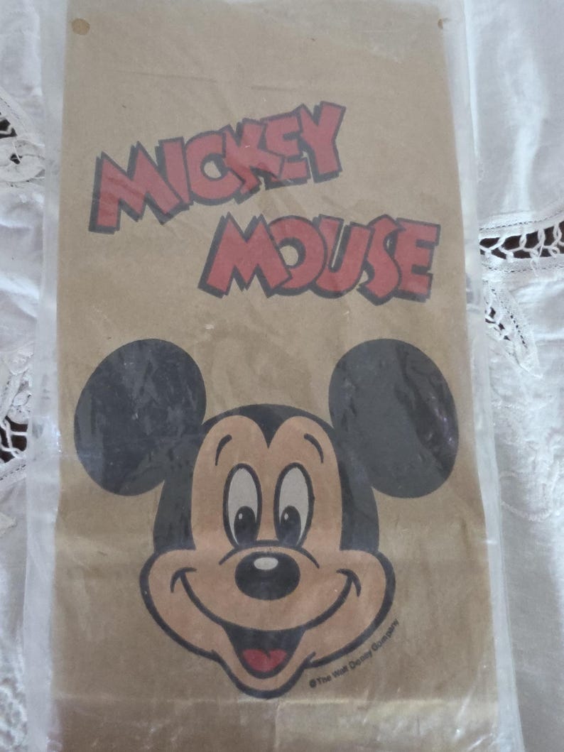 May include: A brown paper bag featuring a smiling Mickey Mouse face and the words "Mickey Mouse" in red letters. The bag has black mouse ears and a red mouth. The text "The Walt Disney Company" is printed at the bottom.
