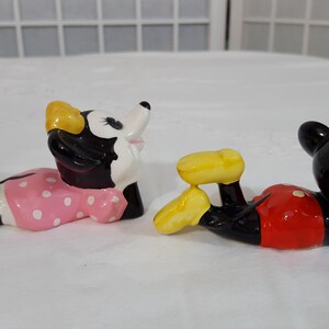 Może przedstawiać: Dwie ceramiczne figurki postaci z kresk&oacute;wek, Myszki Minnie i Myszki Miki, leżą. Myszka Minnie ma na sobie r&oacute;żową sukienkę w białe grochy i niebieskie buty. Myszka Miki ma na sobie czerwone szorty i ż&oacute;łte buty.