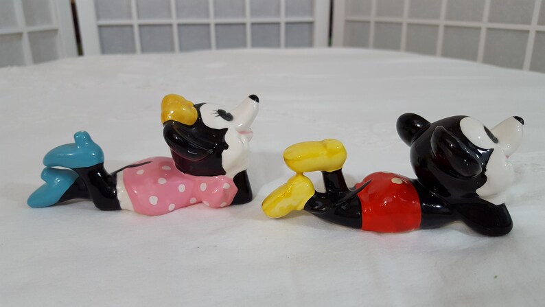 Może przedstawiać: Dwie ceramiczne figurki postaci z kresk&oacute;wek, Myszki Minnie i Myszki Miki, leżą. Myszka Minnie ma na sobie r&oacute;żową sukienkę w białe grochy i niebieskie buty. Myszka Miki ma na sobie czerwone szorty i ż&oacute;łte buty.