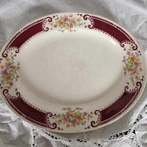 May include: Oval serving platter with a cream-colored surface, a wide burgundy border, and floral accents. The border features decorative scrollwork and small floral bouquets in pink, yellow, and blue. The platter has a gold rim.