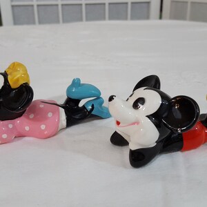 Może przedstawiać: Zestaw trzech ceramicznych figurek przedstawiających Myszki Miki i Minnie. Myszka Minnie ma na sobie r&oacute;żową sukienkę w białe grochy i ż&oacute;łtą kokardę. Miki ma na sobie czerwone szorty i ż&oacute;łte buty. Figurki są w pozycji leżącej.