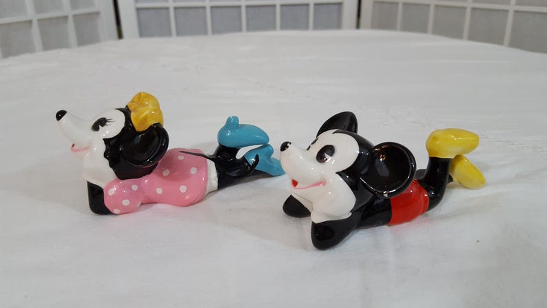 Może przedstawiać: Zestaw trzech ceramicznych figurek przedstawiających Myszki Miki i Minnie. Myszka Minnie ma na sobie r&oacute;żową sukienkę w białe grochy i ż&oacute;łtą kokardę. Miki ma na sobie czerwone szorty i ż&oacute;łte buty. Figurki są w pozycji leżącej.