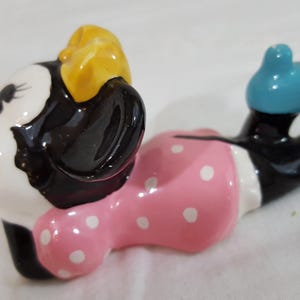 Może przedstawiać: Ceramiczna figurka Myszki Minnie w pozycji leżącej. Figurka ma białą twarz, czarne uszy i ż&oacute;łtą kokardę. Nosi r&oacute;żową sukienkę w białe grochy i niebieskie buty. Jest to przedmiot kolekcjonerski.